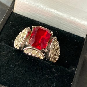 Könnte beinhalten: Ein silberner Ring mit einem großen, rechteckigen roten Edelstein, der von kleineren, klaren Edelsteinen umgeben ist.