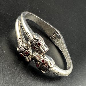 May include: A silver-toned hinged bangle bracelet featuring two stylized animal heads facing each other. The heads have textured surfaces and small, round, deep red stones set as eyes. The metal band has a textured finish and a secure hinge mechanism.