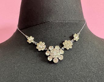 May include: A silver-toned necklace featuring a floral design. The necklace has a delicate chain and multiple flower-shaped pendants, each adorned with sparkling clear crystals. The central flower is larger than the others.