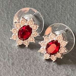 May include: Stud earrings with a central oval, deep red gemstone, encircled by smaller, clear crystals. The earrings have a silver-coloured metal setting and clear plastic backings. A classic jewellery design.