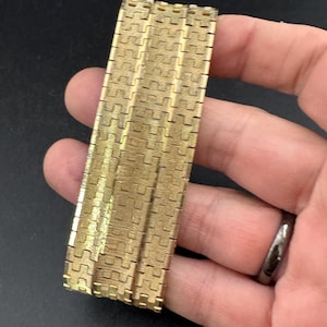 以下が含まれることがあります： 幾何学模様が繰り返されるゴールドトーンのメタルブレスレット。 ブレスレットは、柔軟なバンドを形成する、インターロックされた長方形のリンクで構成されています。 リンクはテクスチャ加工された表面を持ち、ブレスレットはヴィンテージのようです。