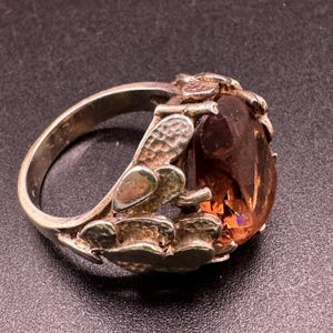 May include: A silver ring featuring a large, oval-cut, amber-coloured gemstone set within a band decorated with textured, leaf-like metal shapes. The metal has a polished finish, and the stone displays a warm, translucent orange hue with facets.