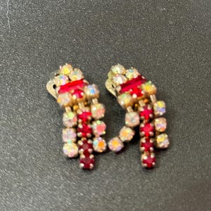 May include: A pair of red and clear rhinestone dangle earrings. The earrings are made of metal and have a clip-on closure.
