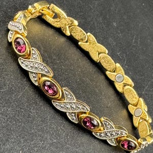 May include: A gold-toned bracelet with a series of oval, magenta-colored gemstones set between X-shaped links. The X-shaped links are accented with small, clear crystals. The bracelet has a clasp closure.