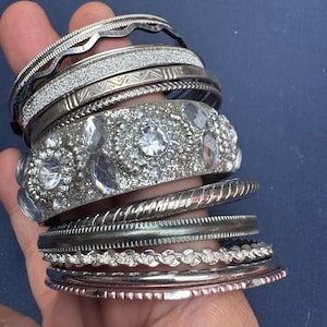 May include: A collection of silver-toned metal bangles and bracelets. The set includes a wide cuff encrusted with clear faceted crystals, several thin bands with textured, glittery, or twisted surfaces, and smooth metallic rings in various widths.