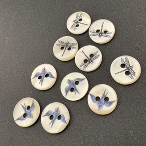 Pu&ograve; includere: Una collezione di bottoni rotondi bianco sporco con due fori. Alcuni bottoni presentano un disegno di libellula nera, mentre altri mostrano un uccello blu. I bottoni sono sparsi su una superficie scura.