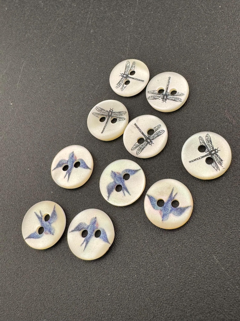 Pu&ograve; includere: Una collezione di bottoni rotondi bianco sporco con due fori. Alcuni bottoni presentano un disegno di libellula nera, mentre altri mostrano un uccello blu. I bottoni sono sparsi su una superficie scura.