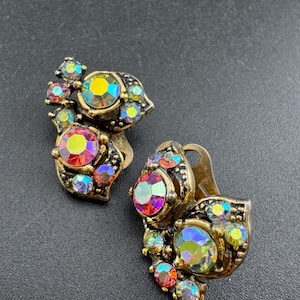 May include: Vintage clip-on earrings with an ornate, gold-tone setting. Each earring features a cluster of iridescent, colorful rhinestones in shades of red, yellow, and blue. The earrings have a classic, elegant design.