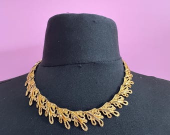 May include: A gold-toned necklace with a delicate, openwork design. The necklace features a repeating pattern of leaf-like shapes, creating a decorative collar. The jewelry is displayed on a black mannequin head against a pink background.