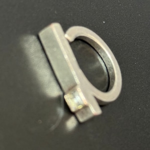 May include: A silver ring with a rectangular band and a small, clear stone set in the centre.