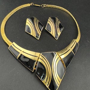 May include: A vintage jewelry set featuring a gold-tone necklace and matching earrings. The necklace has a geometric design with black enamel accents. The earrings are diamond-shaped, and the set is displayed on a dark surface.