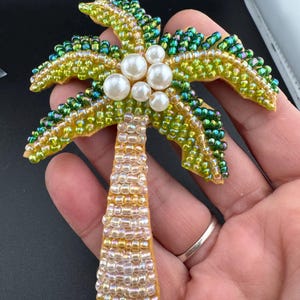 May include: A beaded palm tree brooch with a detailed design. The brooch has a trunk in shades of gold and a crown of green and yellow beads. Five white pearl beads are clustered in the center of the palm fronds. The brooch is held in a hand.