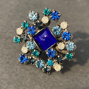 May include: A silver brooch featuring a blue square stone surrounded by smaller blue, light blue, and white rhinestones.