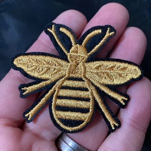 May include: A gold embroidered bee patch with black outlining. The bee has a striped abdomen and is shown in profile.
