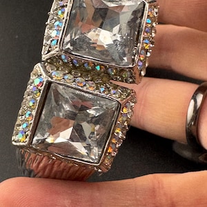 以下が含まれることがあります： 2つの大きな正方形の透明な宝石が付いた、シルバーカラーのブレスレット。ブレスレットは、小さな虹色のラインストーンで装飾されています。大胆なジュエリーです。