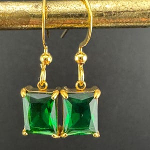 May include: A pair of gold-colored earrings with emerald-green rectangular gemstones. The earrings have a simple design with a hook closure and a small gold bead above the gemstone. The gemstones are set in a gold frame.