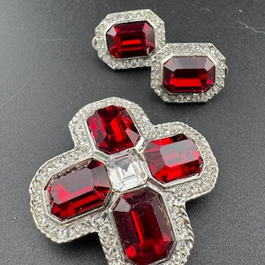 May include: A silver-tone brooch with a cross design featuring five red and one clear faceted stones surrounded by smaller clear stones. A matching pair of earrings with red faceted stones are also included.