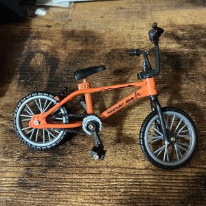 Op de afbeelding: Een kleine oranje mountainbike met zwarte onderdelen en grijze wielen. Op het frame staat de tekst "MOUNTAIN BIKE". Deze miniatuurfiets is waarschijnlijk een speelgoed of een decoratief item.