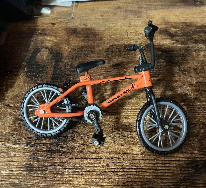 Op de afbeelding: Een kleine oranje mountainbike met zwarte onderdelen en grijze wielen. Op het frame staat de tekst "MOUNTAIN BIKE". Deze miniatuurfiets is waarschijnlijk een speelgoed of een decoratief item.