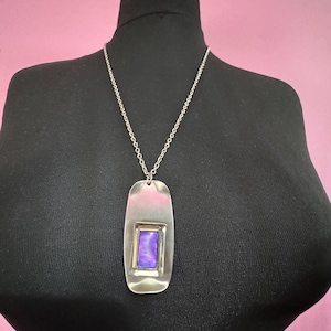 May include: A silver-toned pendant necklace with a rectangular purple stone set in the center. The pendant is a long, rectangular shape with rounded edges and hangs from a delicate chain. The necklace is displayed on a black mannequin.