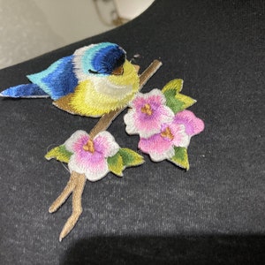May include: Embroidered iron-on patch featuring a blue and yellow bird perched on a branch with pink flowers.