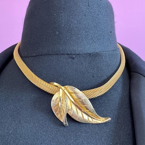 Könnte beinhalten: Goldfarbene Halskette mit einem Mesh-Kettenglied und einem Blattanhänger. Der Blattanhänger hat detaillierte Adern und eine strukturierte Oberfläche. Die Halskette wird auf einem schwarzen Hintergrund mit einem violetten Hintergrund präsentiert.