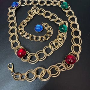 Könnte beinhalten: Ein goldener Kettengürtel mit mehreren großen, runden, cabochonförmigen Edelsteinen in verschiedenen Farben. Die Edelsteine sind rot, grün und blau.