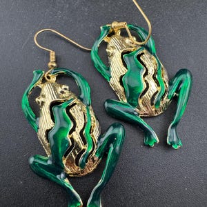 May include: A pair of frog-shaped earrings. The earrings have a gold-coloured base with green enamel detailing. The frog's body has a textured design, and the legs are extended. The earrings have gold-coloured ear wires.