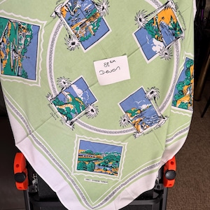 May include: A light green and white square tablecloth featuring framed images of coastal scenes with palm trees and the text "Torquay Harbour" and "Anstey's Cove". The tablecloth has a white border and a decorative edge.