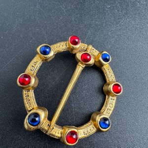 May include: A gold-toned brooch with a circular design, adorned with alternating red and blue gemstones. The brooch features a pin closure and intricate detailing around the edges, creating a vintage aesthetic.
