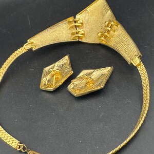 May include: A gold-toned necklace and matching earrings set. The necklace features a wide, textured collar with a geometric design and a chain. The earrings are diamond-shaped with a textured surface. The jewelry set is displayed on a dark surface.