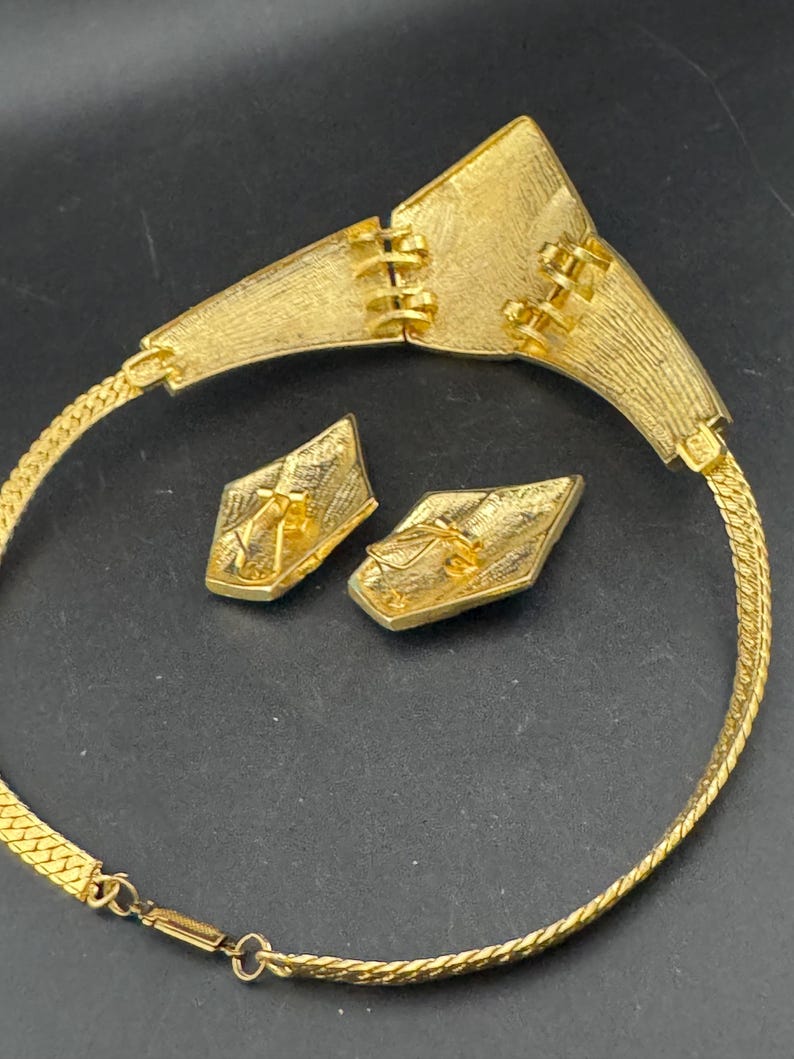 May include: A gold-toned necklace and matching earrings set. The necklace features a wide, textured collar with a geometric design and a chain. The earrings are diamond-shaped with a textured surface. The jewelry set is displayed on a dark surface.