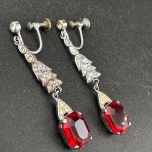 May include: A pair of vintage silver-toned clip-on earrings. Each earring features a decorative top with clear rhinestones, a section of small yellow stones, and a large, rectangular, faceted, red gemstone pendant. The earrings are displayed on a dark surface.