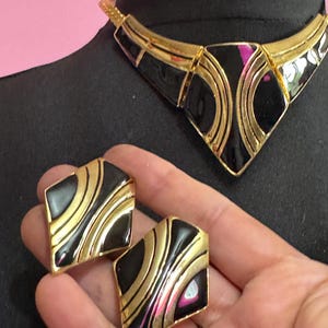 May include: A vintage jewelry set featuring a black and gold necklace and matching earrings. The necklace has a geometric design with black enamel and gold-tone metal. The earrings are square-shaped with a similar black and gold pattern. The set is displayed on a black mannequin.