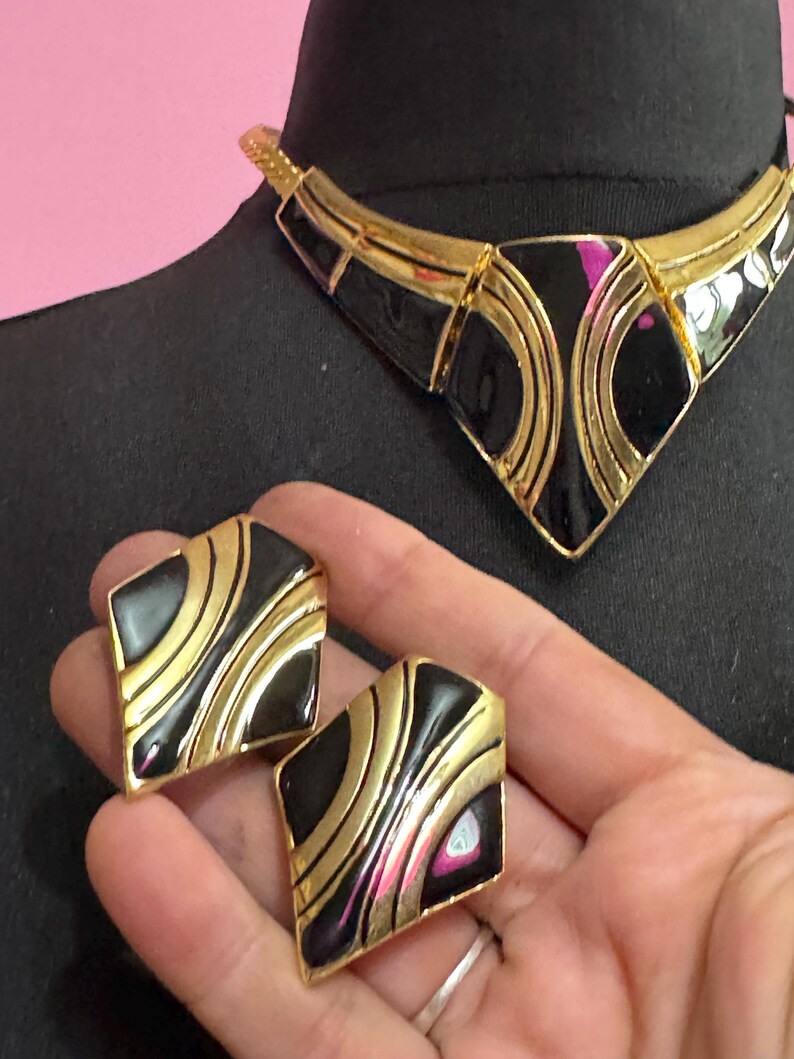 May include: A vintage jewelry set featuring a black and gold necklace and matching earrings. The necklace has a geometric design with black enamel and gold-tone metal. The earrings are square-shaped with a similar black and gold pattern. The set is displayed on a black mannequin.