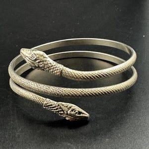 May include: A silver-toned metal wrap bracelet shaped like a coiled snake. The design features two detailed snake heads at each end of the spiral band, with a textured surface that mimics snake scales covering the entire length of the metal coil.