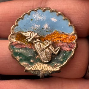 May include: A vintage, round pin with a scalloped edge. The pin features a silver figure of a seated elf against a backdrop of a blue sky with white clouds, orange and brown mountains, and green and pink ground. The pin has a gold-colored metal border.