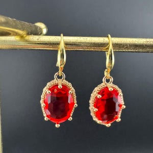 May include: A pair of gold earrings featuring red, oval gemstones. The gems are set in a decorative gold frame. The earrings hang from gold hooks. The gemstones are approximately 1.5 cm in length.