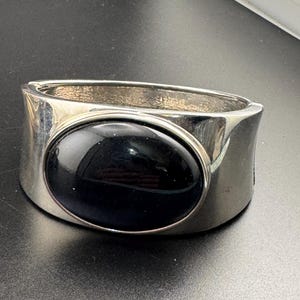 May include: Silver-tone cuff bracelet with a large, oval black stone. The bracelet has a wide band with a polished finish. The black stone is set in a bezel setting, creating a striking contrast.