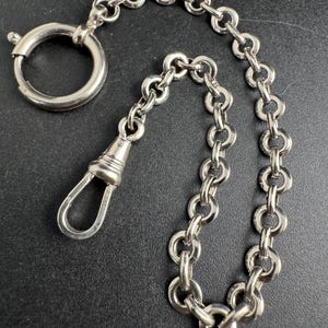May include: A silver pocket watch chain featuring a spring ring clasp and a swivel clasp. The chain is made of interconnected oval links. The background is a dark, solid colour.