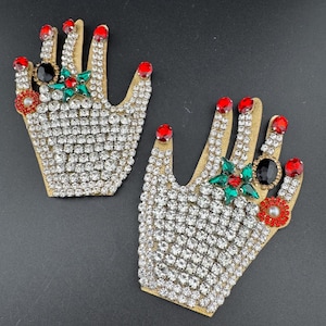 May include: Two decorative hand-shaped patches. Each patch is covered in clear rhinestones and features red nail polish, a black gem ring, and a green and red gem design. The hands are outlined in gold.