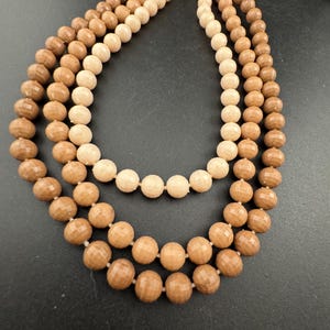 May include: A multi-strand necklace featuring faceted, round beads in shades of brown and cream. The necklace has three strands of varying lengths, creating a layered effect. The beads are strung on a delicate chain with a gold-tone clasp.