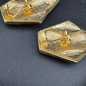 May include: Close-up of two gold-toned, geometric earrings. Each earring has a textured surface and a post-and-clip closure. The earrings are set against a dark background, highlighting their metallic sheen and intricate design.