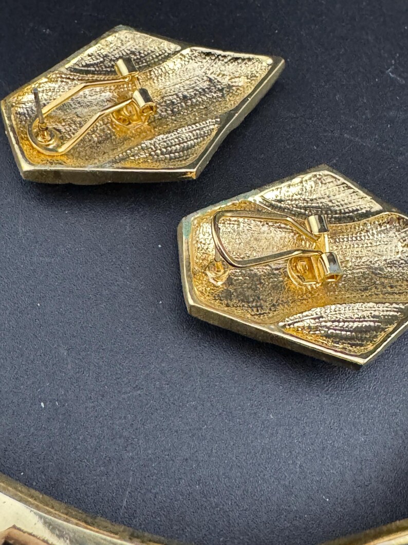 May include: Close-up of two gold-toned, geometric earrings. Each earring has a textured surface and a post-and-clip closure. The earrings are set against a dark background, highlighting their metallic sheen and intricate design.