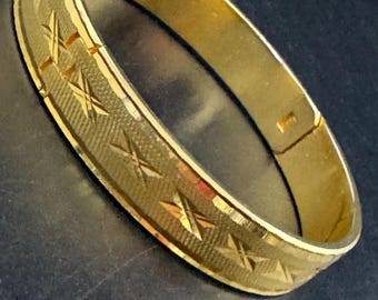 Peut inclure: Un bracelet jonc doré. Le bracelet présente une bande texturée avec un motif d'éclat d'étoile répété. Le fermoir est visible sur le côté. Le bijou est présenté sur une surface sombre.