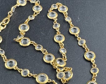 May include: A gold-toned necklace featuring clear, round gemstones set in individual bezels. The gemstones vary in size and are linked by a delicate chain. The necklace is displayed against a dark background.