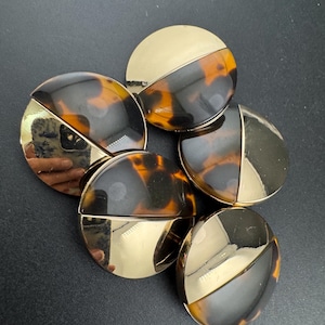 May include: Six large, round buttons with a two-tone design. Half of each button is gold, and the other half features a tortoiseshell pattern in brown and black. The buttons are displayed on a dark surface. These buttons could be used for clothing.