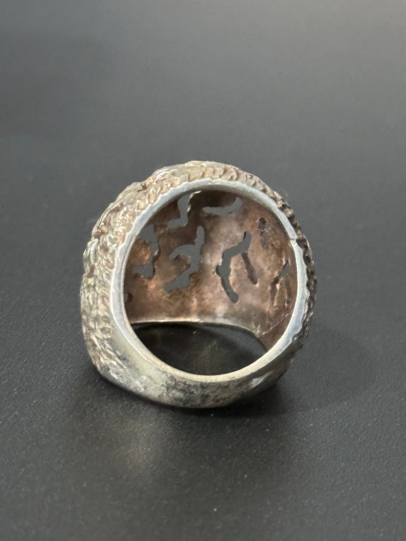 May include: A silver ring with an intricate, openwork design. The ring is made of a textured metal and has a wide band.