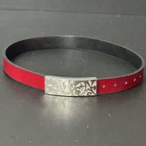 May include: A red leather bracelet with a silver buckle featuring a floral design.