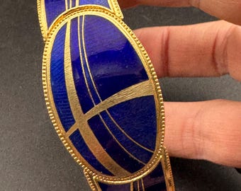 May include: A gold-toned bracelet with a striking design. The bracelet features oval-shaped links with a deep blue enamel finish, accented by gold lines. The edges are detailed with a delicate beaded pattern, creating an elegant accessory.